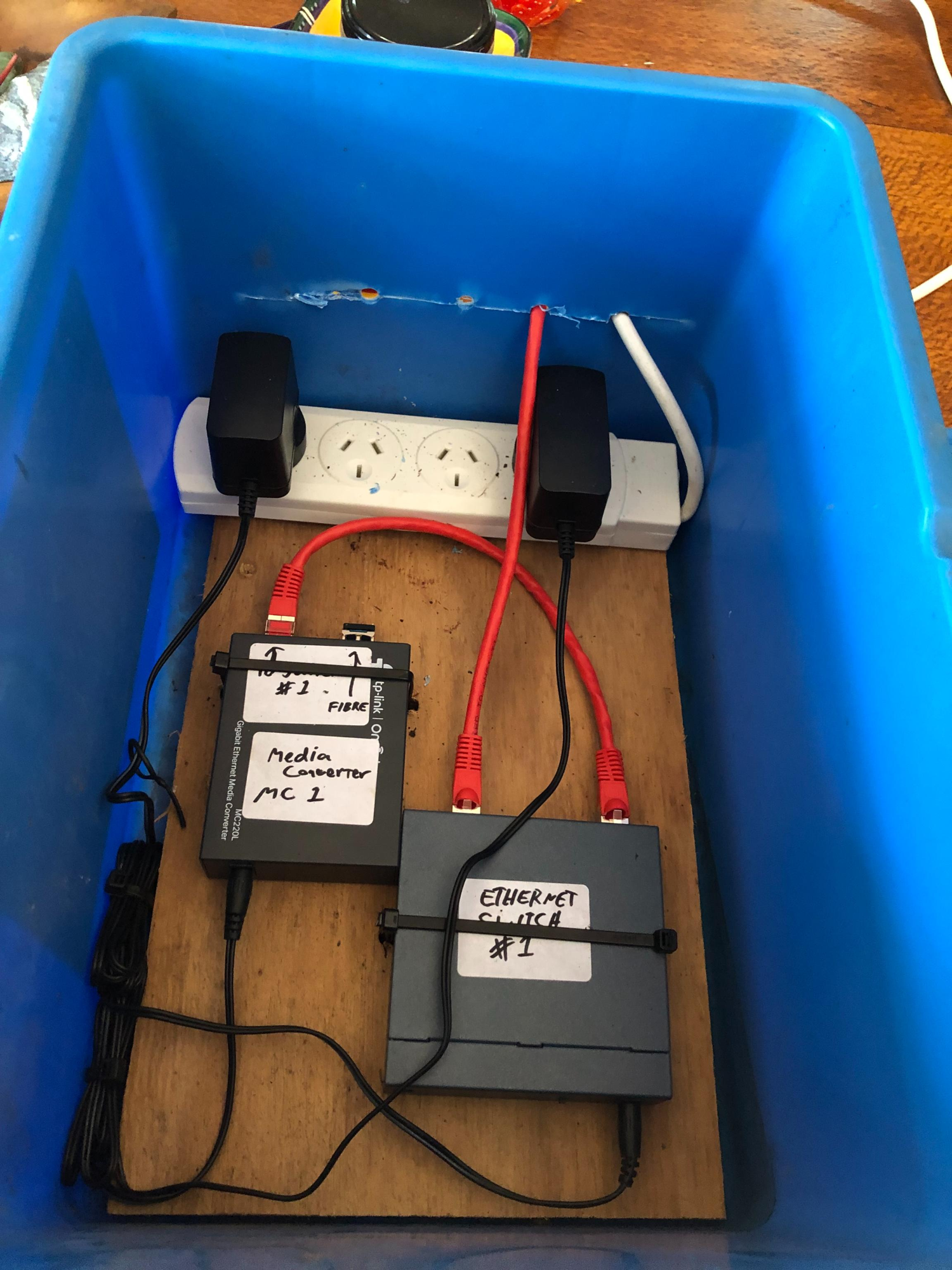 The inside of a blue plastic tub mounted as an outdoor enclosure. A TP-Link MC220L gigabit media converter labelled 'Media Converter MC 1' sits next to an Ethernet switch labelled 'Ethernet Switch #1', both wired to a small power board with two adapters.
