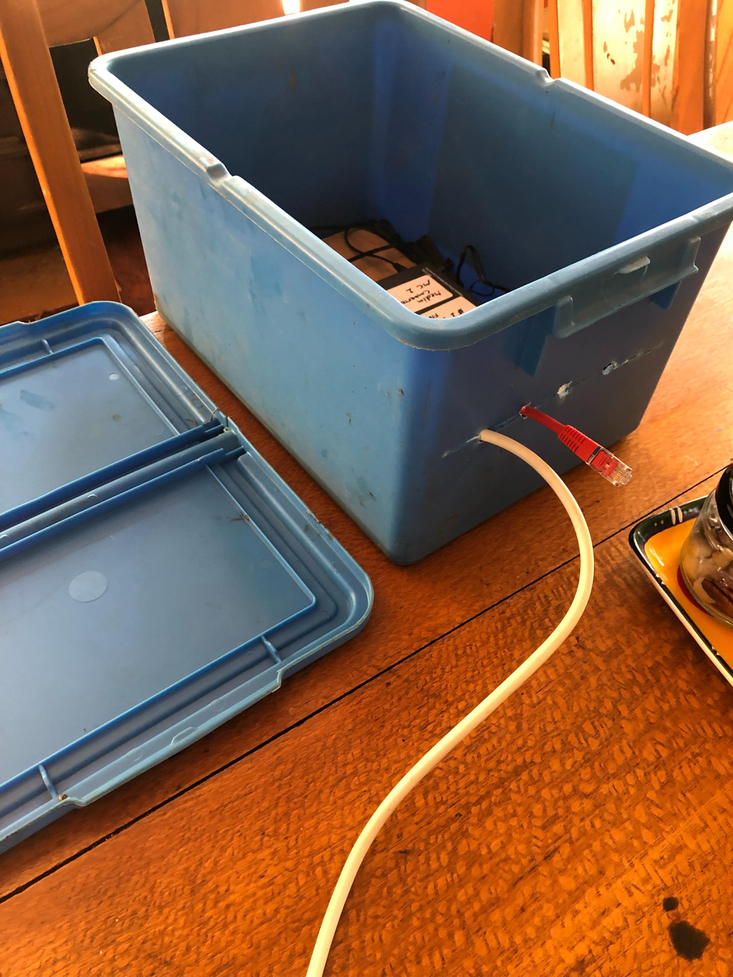The outside of the blue plastic tub on a wooden table, lid set aside, with a red Ethernet cable and a white fibre cable feeding in through a drilled hole on the side.