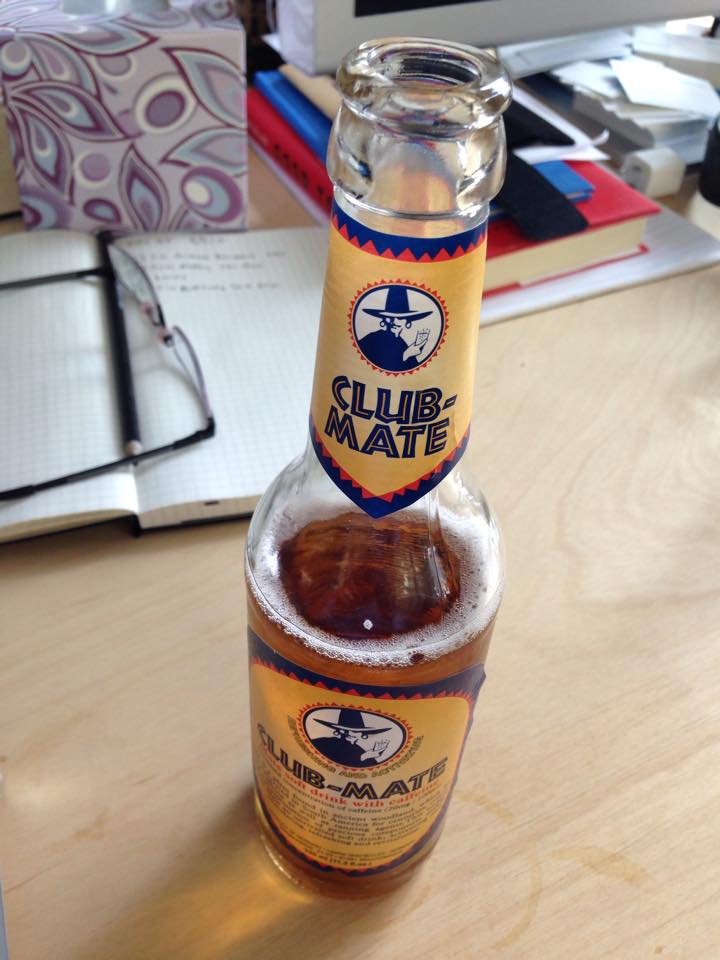 A half-drunk bottle of Club-Mate on a wooden desk, next to an open grid notebook, a pair of glasses and a stack of books.