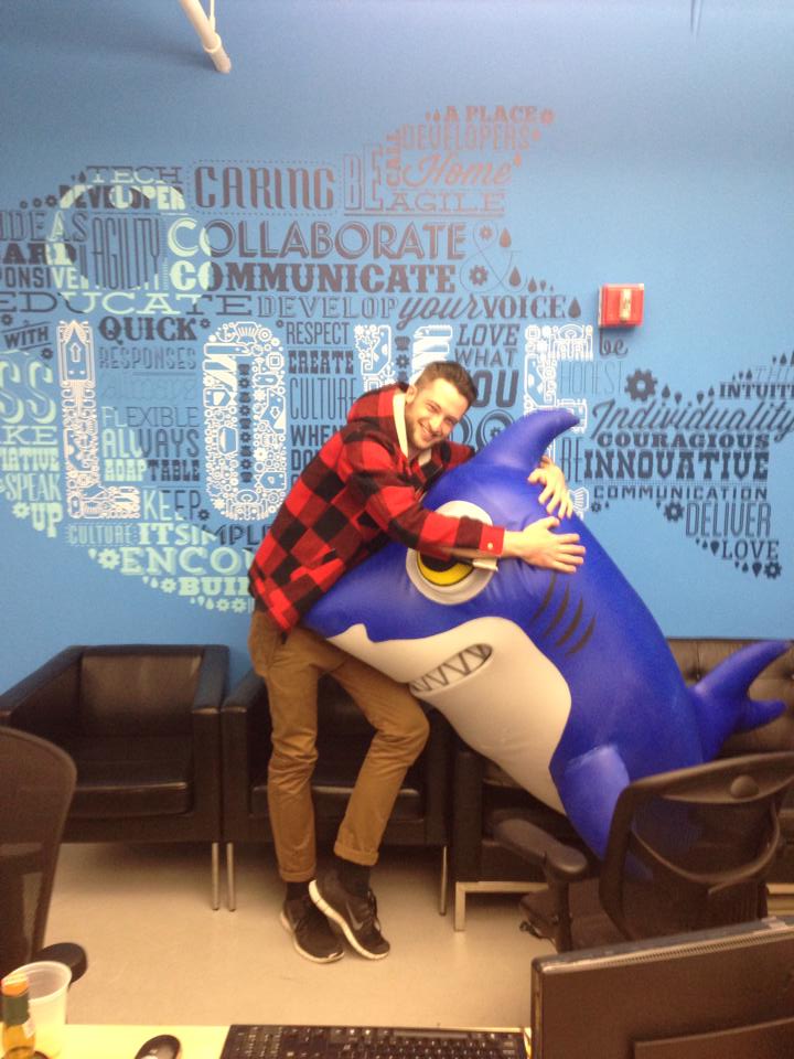 Ivan in a red plaid flannel shirt hugging a giant inflatable blue shark in front of a blue wall covered in words like Collaborate, Communicate and Innovative.